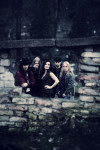 Nightwish-bändin jäsenet seisovat tummasävyisessä ja taiteellisessa valokuvassa keskellä ruhjoutuneen kivimuurin tai kiviseinän takaa. Taustalla vanha kivirakennus, joka on rikki.