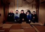 Viisi Radiohead-bändin miestä istuu rivissä seinää vasten. Huone on täynnä laattamaisia nahkasuikaleita, jotka peittävät lattian ja seinät. Miehillä yllään arkivaatteet.