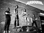 Mustavalkoinen bändivalokuva Airbourne-yhtyeen jäsenistä, jotka seisovat auton päällä. Taustalla suuri tiiliseinä. Miehiä kuvassa neljä kappaletta. Heistä jokaisella pörröiset hiukset.