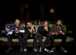 Valokuva Coldplayn kokoonpanoon kuuluvista neljästä miehestä, jotka istuvat sohvalla rivissä. Vasemmanpuoleinen mies soittaa sinivalkoista kitaraa ja muut katselevat kierosti ylös.