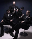 Valokuva neljästä CMX-bändin miesjäsenestä, joista kaksi etummaista istuu mustilla tuoleilal samalal kun kaksi muuta seisoo takana tummanviolettia taustaa vasten. Miehillä yllään tummat puvut, osalla myös lierilakit päässä.