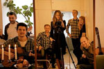Scandinavian Music Groupin jäsenet istuvat ja seisovat tiiviinä ryhmänä oleskeluhuoneessa. Kuva on väritykseltään lämpimänkeltainen. Vasemmalla kruunukynttilöitä. Kuvassa miehiä ja yksi nainen, joka mustiin pukeutuneena seisoo heidän keskellään.