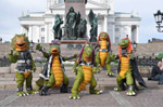 Valokuav suuriin dinosaurusasuihin pukeutuneista Hevisauruksen jäsenistä, joita on kuvassa yhteensä viisi kappaletta. Dinot kävelevät kaksin jaloin ja ne seisovat ulkoilmassa koristeellisten patsaiden edessä torilla.