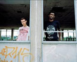 Massive Attackin kaksi jäsentä, jotka ovat miehiä, seisovat harmaan betonimaisen rakennelman sisällä suuren betonipylvään toisilla puolilla. Vasemmanpuoleinen on valkoihoinen ja valkopaitainen, oikeanpuoleinen mustaihoinen ja mustapaitainen.