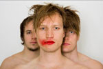 Valokuva kolmesta lyhythiuksisesta miehestä seisomassa valkoista taustaa vasten siten, että keskimmäisenä on yksi mies, jonka takana kaksi muuta heppua seisovat miehen takaraivon takana. Keskimmäisellä miehellä punaista huulipunaa.