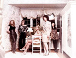 Ylivalottunut ryhmäkuva Viisi-yhtyeen naisista. Naisia yhteensä viisi kappaletta, joista osa seisoo seiniä vasten ja keskimmäinen istuu valkoisella tuolilla.